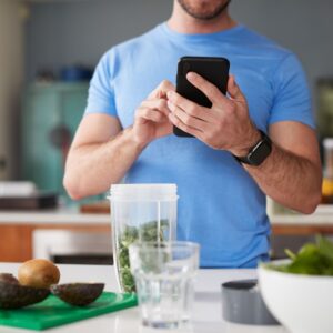 Man using a fitness/calorie tracker app on his smartphone in the kitchen to monitor nutrition and track progress.